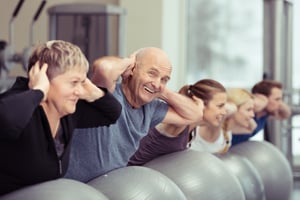 Elderly couple doing pilates class at the gym with a group of diverse younger people balancing on the gym ball with raised arms to tone their muscles in an active retirement concept-1